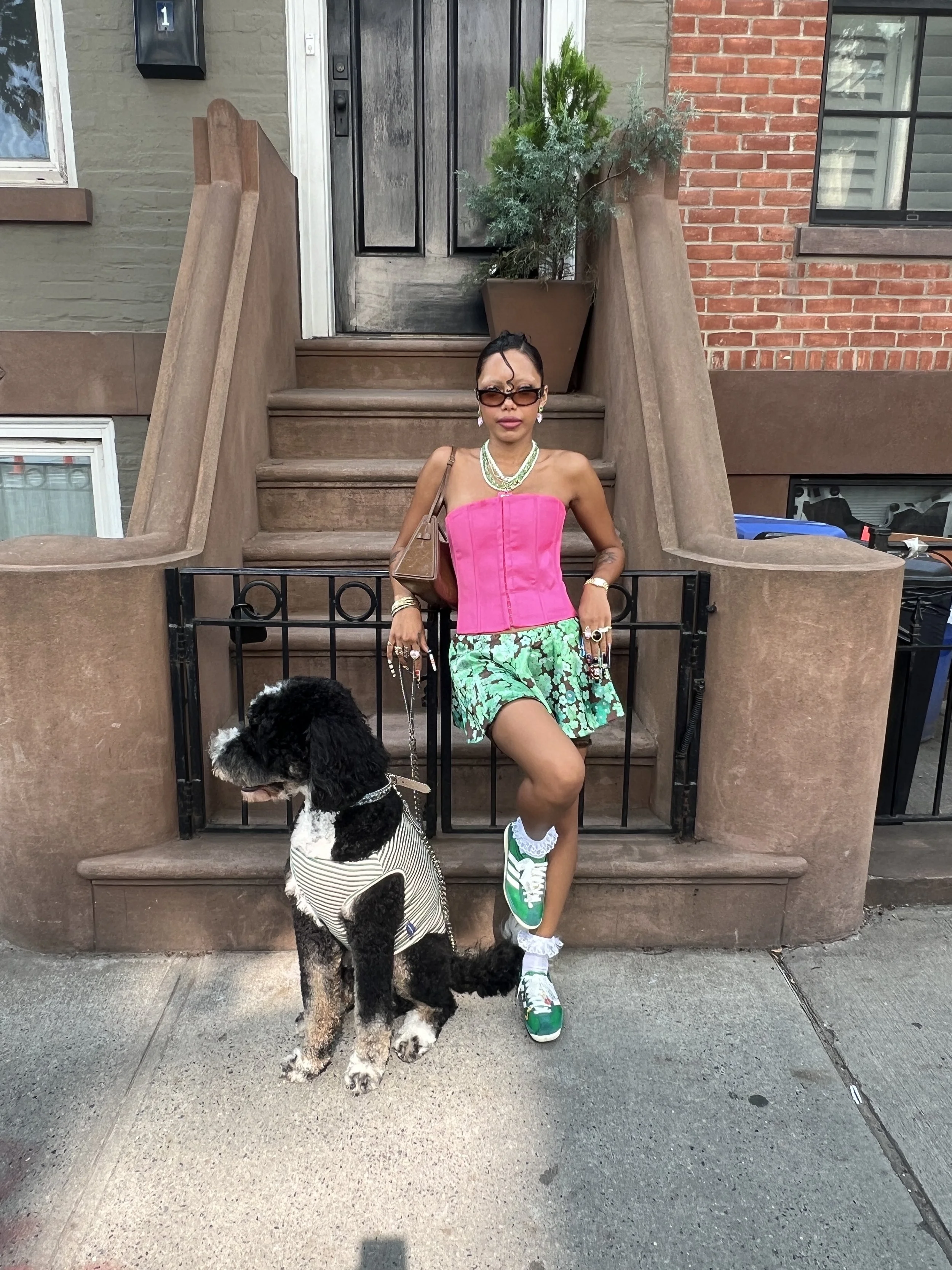 A woman dressed in a pink corset, green skirt, green sneakers, and sunglasses stands on a stoop with her black and white dog wearing a striped outfit. They are outside a building with brick and painted walls.