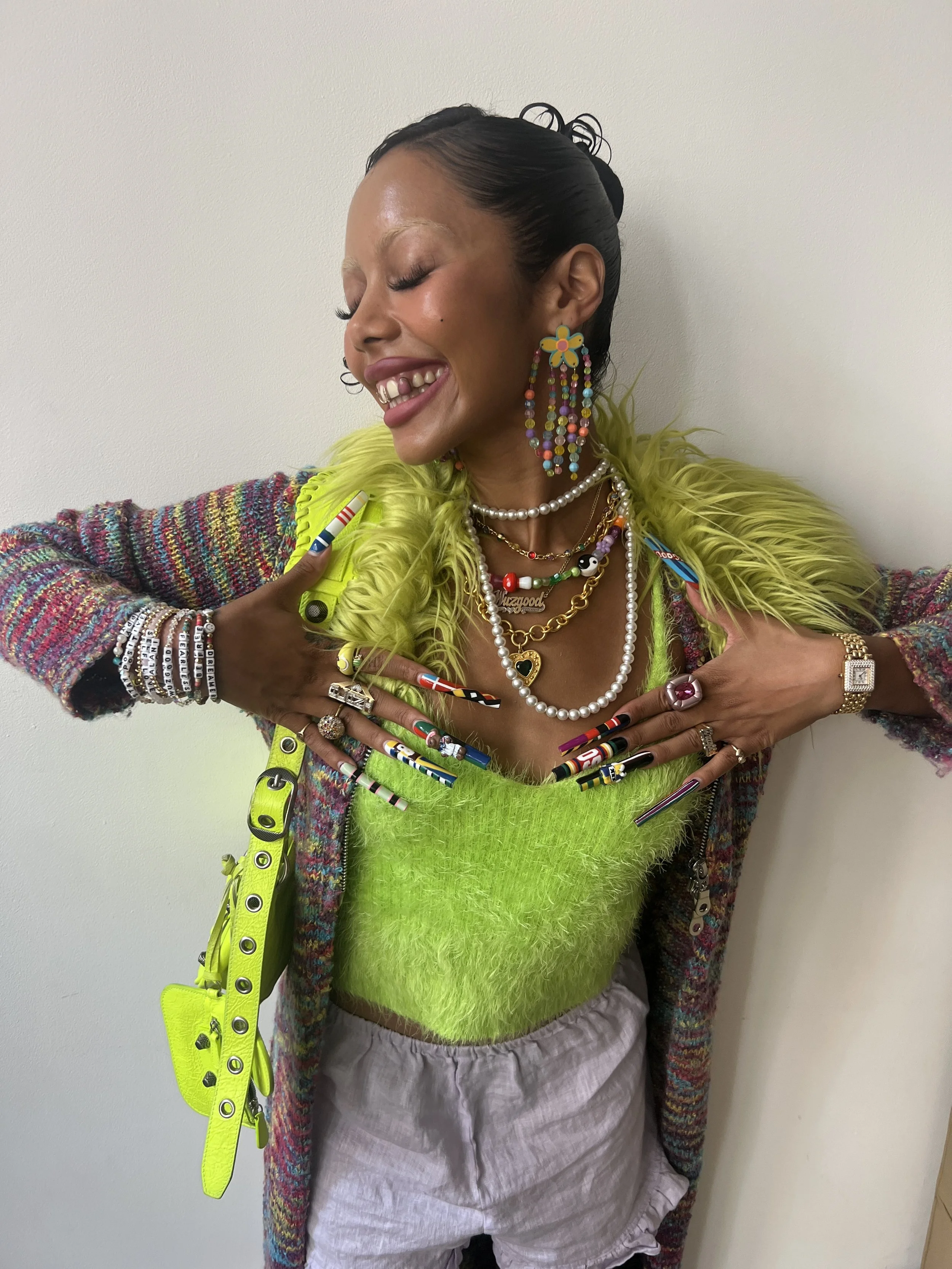 Woman with colorful, layered jewelry, neon green fuzzy top, and multicolored nail art, smiling with eyes closed and arms crossed in front of her chest, wearing a rainbow-colored jacket and beige shorts.