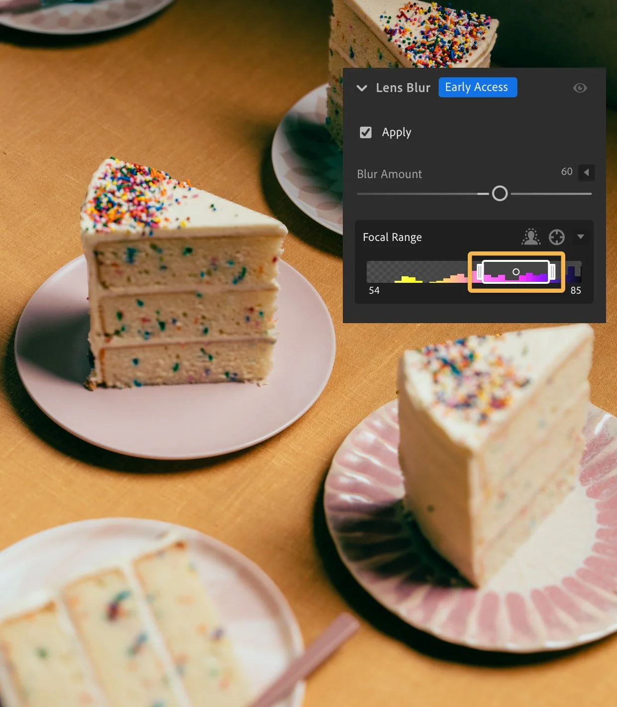 Slices of birthday cake with white frosting and colorful sprinkles on pink and white plates, on a wooden table, with a photo editing overlay showing lens blur settings a feature in Adobe Lightroom.