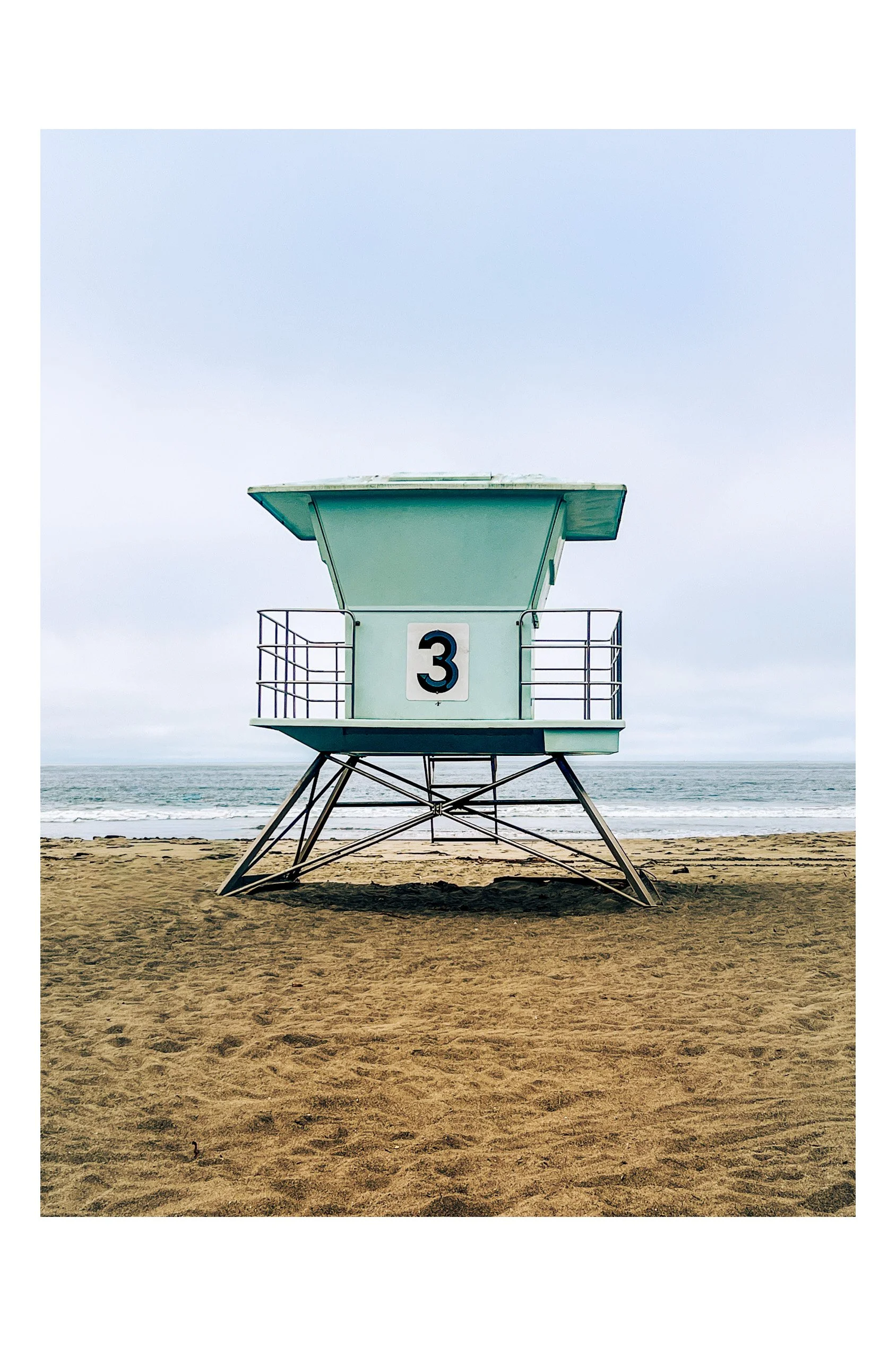A lifeguard tower with the number 3, standing on the sandy beach beside the ocean under a cloudy sky. California beach scene.