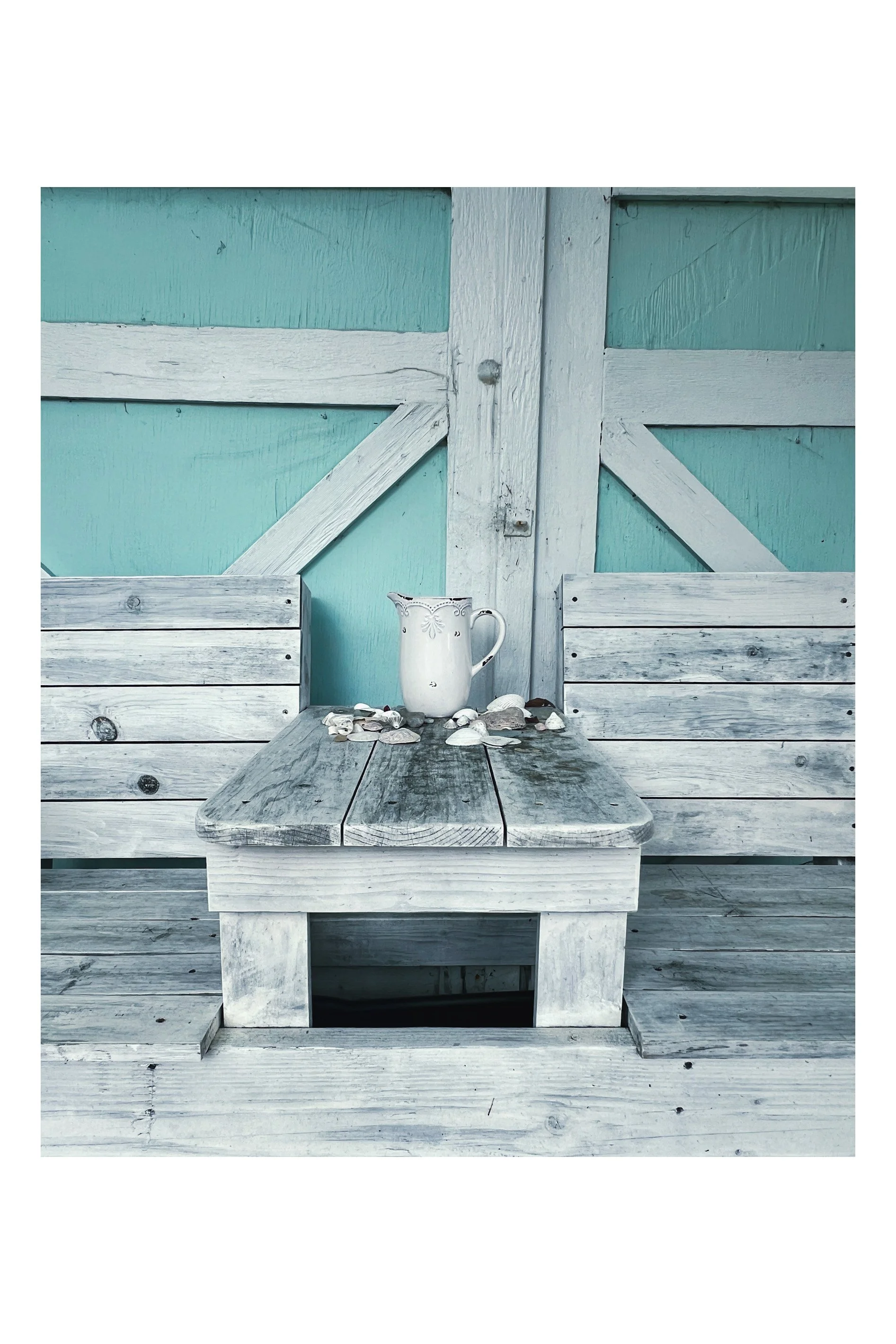 A weathered wooden table with a white pitcher surrounded by seashells, set against a rustic turquoise and white wooden background.